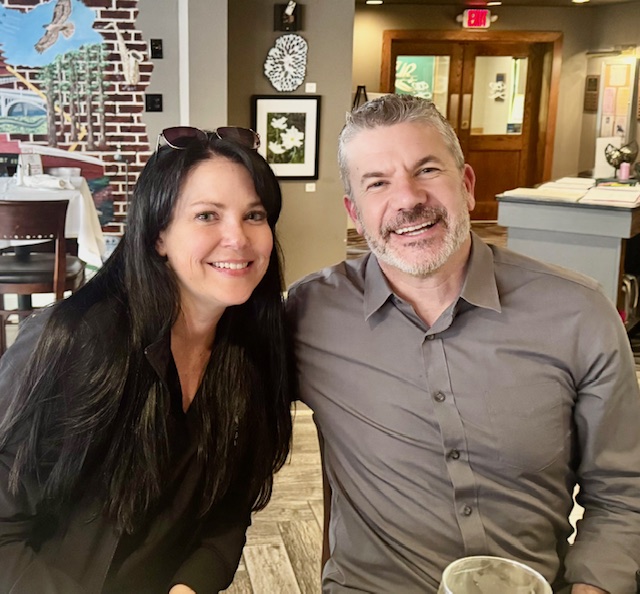 Two people stand close together and pose inside a restaurant with brick walls, framed artwork, and a mural in the background.