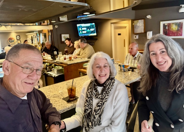 Three people stand close together and pose inside a restaurant with brick walls, framed artwork, and a mural in the background.