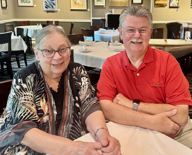 Two people sit close together and pose inside a restaurant with brick walls, framed artwork, and a mural in the background.