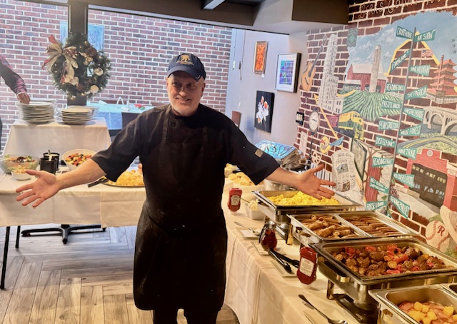 A man in black attire poses in front of the camera. Behind him are tables with an assortment of foods.