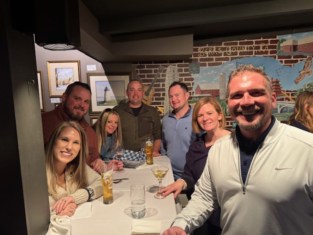 A group of people are gathered around a table in a restaurant setting with drinks, food, framed artwork, and a mural visible in the background.