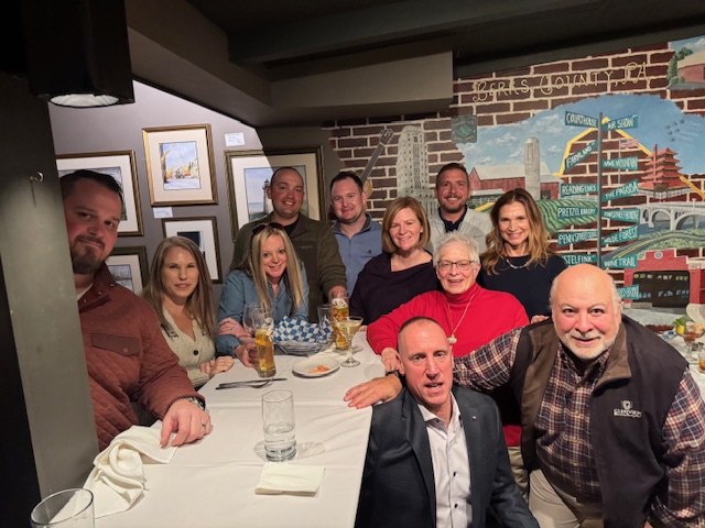 A group of people are gathered around a table in a restaurant setting with drinks, food, framed artwork, and a mural visible in the background.